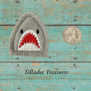 May include: A gray felt shark head with a red and white embroidered mouth and black eyes. A U.S. quarter coin is next to the shark head. The text "Telladee Treasures" is at the bottom.