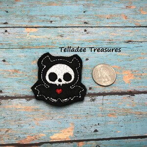 May include: A black felt embroidered patch with a white skull and red heart, shaped like a bat. The patch is about the size of a US quarter.