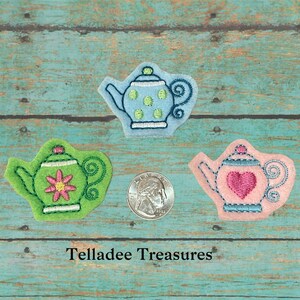 May include: Three embroidered felt teapots in different colors and designs. The blue teapot has white polka dots, the green teapot has a pink flower, and the pink teapot has a red heart.