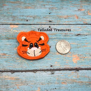 May include: Orange felt tiger face applique with black stripes, white nose, and pink eyes. The applique is about the size of a US quarter.