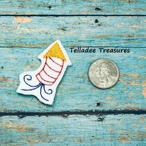 May include: A white felt embroidered rocket with red and white stripes, a yellow pointed top, and white and blue tails. The rocket is about the size of a US quarter.