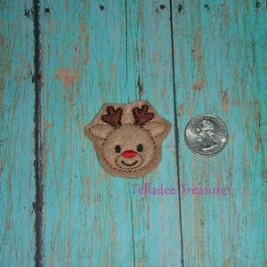 May include: A tan felt reindeer head with brown antlers and a red nose. The reindeer is next to a U.S. quarter coin. The background is a weathered, turquoise-colored wooden surface. The text "Telladee Treasures" is in the lower left corner.