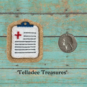 May include: A beige felt keychain with a white and black embroidered image of a medical clipboard with a red cross. The keychain is about the size of a quarter.