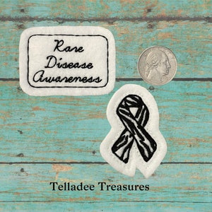 May include: Two white felt patches with black embroidery. One patch features a zebra-striped ribbon and the other says "Rare Disease Awareness".