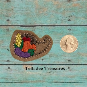 May include: A small, brown felt applique in the shape of a cornucopia filled with embroidered autumn harvest vegetables. The applique is about the size of a US quarter.