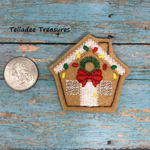 May include: A felt gingerbread house ornament with a red bow, green wreath, and yellow lights. The ornament is approximately the size of a US quarter.
