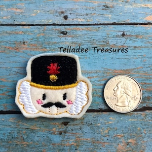 May include: A white felt nutcracker ornament with a black hat, gold trim, red feather, and a black mustache. The ornament is about the size of a US quarter.