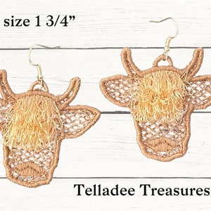 May include: A pair of brown lace cow earrings with gold fringe. The earrings are 1 3/4 inches in size. The text "Telladee Treasures" is visible below the earrings.