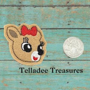 May include: A brown felt applique of a reindeer face with a red bow, black eyes, and a white nose. The applique is about the size of a US quarter.