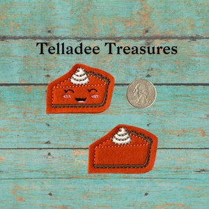 May include: Two felt pumpkin pie slices with smiling faces and whipped cream. The pie slices are brown with a light brown crust and white whipped cream. The pie slices are about the size of a US quarter.