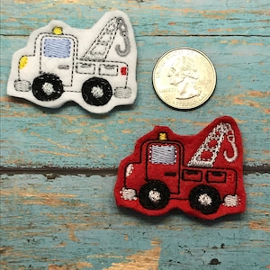 May include: Two embroidered felt tow truck patches. One is white with a yellow stripe and a blue window. The other is red with a blue window. Both have a gray tow arm and hook.