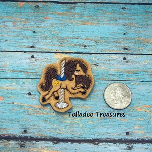 May include: A tan felt carousel horse patch with brown mane and tail, a blue bow, and a yellow flower. The patch is on a blue wooden surface. The text "Telladee Treasures" is below the patch.