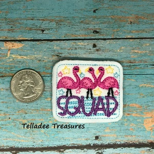 May include: Embroidered iron-on patch with three pink flamingos and the word "Squad" in purple letters on a blue and white striped background. The patch is surrounded by yellow stars.
