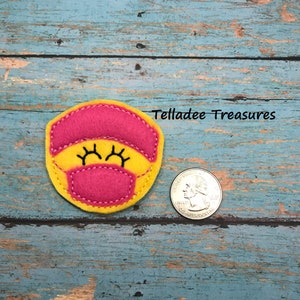 May include: A yellow felt appliqué with a pink face mask and closed eyes. The appliqué is about the size of a US quarter.