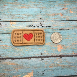 May include: A brown felt patch shaped like a bandage with a red embroidered heart in the center. The patch is about the size of a US quarter.