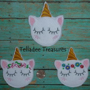 May include: Three white felt unicorn head appliques with gold horns. The top applique has closed eyes. The middle applique has closed eyes and green and pink floral embroidery. The bottom applique has closed eyes and blue, pink, and yellow floral embroidery. The text "Telladee Treasures" is visible below the top applique.