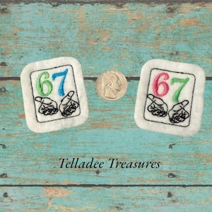 May include: Two white embroidered patches with the number 67 in green and blue, and red and green, with hand gestures. A quarter is next to the patches. The patches are on a teal wooden surface. The text "Telladee Treasures" is at the bottom.
