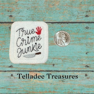 May include: White felt square patch with black embroidery that reads "True Crime Junkie" with a red handprint and a black knife graphic.