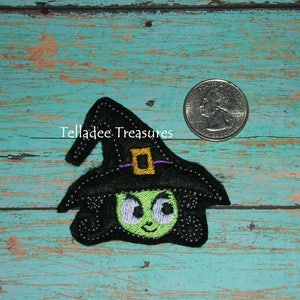 May include: A black witch applique with a green face, large eyes, and a black hat with a gold buckle and purple strap. The applique is on a teal wooden surface. The text "Telladee Treasures" is visible.