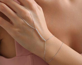 May include: A delicate silver hand chain bracelet. The bracelet features a bar of small, sparkling stones across the back of the hand, connected by fine chains to the wrist and a finger ring. The jewelry is displayed on a person's hand.