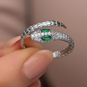 Può includere: Un anello a forma di serpente argentato con corpo strutturato e testa con due gemme verdi ovali. L'anello è incastonato con piccole gemme trasparenti. La testa del serpente si curva attorno al dito.