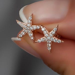 May include: Rose gold starfish stud earrings encrusted with sparkling clear crystals. The earrings are held up by a person's hand. The earrings are a beach-themed jewelry item.
