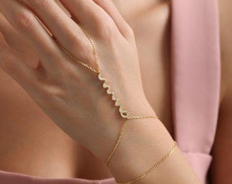 Op de afbeelding: Een goudkleurige handketting met de naam "Anna" uitgeschreven in kleine, sprankelende kristallen. Het delicate sieraad verbindt met een armband met een fijne ketting. De handketting wordt gedragen op de hand van een persoon.