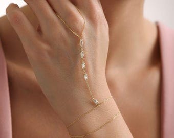 Op de afbeelding: Een delicate gouden handkettingarmband met een reeks rechthoekige, heldere edelstenen. De armband verbindt met een polsketting met twee strengen. De sieraden zijn ontworpen om op de hand en pols te worden gedragen.