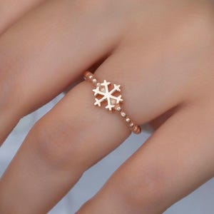 Puede incluir: Anillo de copo de nieve de oro rosa. El anillo presenta un delicado diseño de copo de nieve en el centro, con pequeñas cuentas redondas a lo largo de la banda. El copo de nieve tiene un patrón calado detallado. El anillo se muestra en un dedo.
