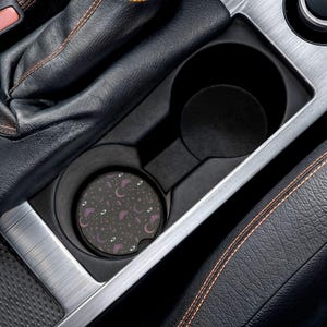May include: A black car coaster with a pattern of purple moons, stars, eyes, and butterflies. The coaster sits in a car's cup holder, surrounded by black leather and silver trim.