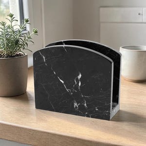 May include: A black marble napkin holder with white veining. The holder has a curved top and a rectangular base. It is sitting on a wooden surface. A small potted plant is to the left.
