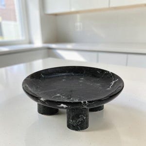 May include: A circular black marble tray featuring natural white veining throughout the stone surface. The tray is elevated by three short, cylindrical legs made from the same black marble material, creating a stable and raised display platform.