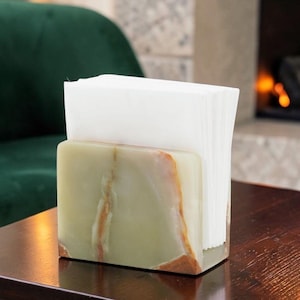 May include: A rectangular napkin holder crafted from polished green onyx stone with natural brown veining patterns. The holder features a smooth, solid base and upright sides that securely contain a stack of folded white paper napkins.