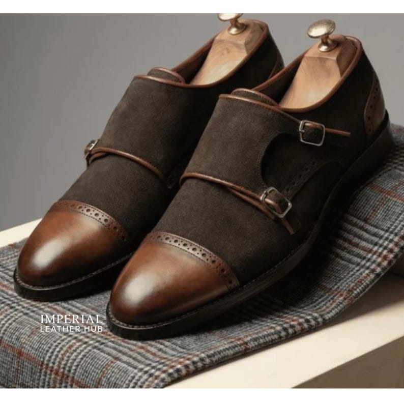 May include: A pair of brown leather double monk strap shoes with a suede upper. The shoes have a cap toe design and are displayed on a patterned fabric. The shoes have a buckle closure and are made of leather. The text "IMPERIAL LEATHER HUB" is visible.