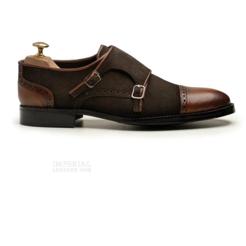 May include: Brown suede double monk strap shoe with leather accents. The shoe features a cap toe, two buckle closures, and a stacked heel. The shoe has a shoe tree inside. The sole is black.