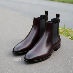 Pu&ograve; includere: Un paio di stivali Chelsea in pelle bordeaux lucida. Gli stivali hanno una finitura elegante e lucida e pannelli laterali elastici. L'immagine mostra gli stivali da una prospettiva bassa, evidenziando la loro forma e il loro design. Gli stivali sono esposti su una superficie grigia.