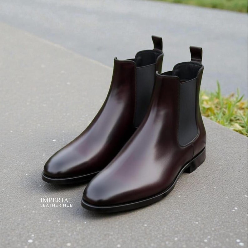 Pu&ograve; includere: Un paio di stivali Chelsea in pelle bordeaux lucida. Gli stivali hanno una finitura elegante e lucida e pannelli laterali elastici. L'immagine mostra gli stivali da una prospettiva bassa, evidenziando la loro forma e il loro design. Gli stivali sono esposti su una superficie grigia.