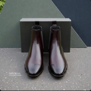 Pu&ograve; includere: Un paio di stivaletti Chelsea in pelle bordeaux lucida. Gli stivaletti hanno un design elegante a punta e pannelli laterali elastici. Sono esposti davanti a una scatola grigio scuro e uno sfondo di forme geometriche verdi e blu navy. Il marchio "IMPERIAL LEATHER HUB" &egrave; visibile.