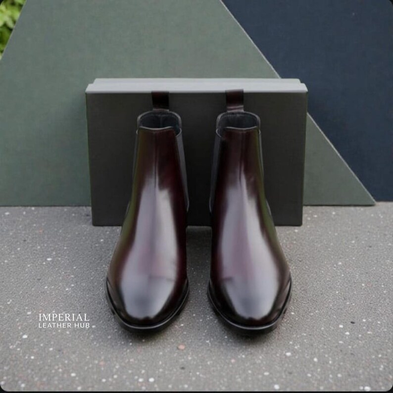 Pu&ograve; includere: Un paio di stivaletti Chelsea in pelle bordeaux lucida. Gli stivaletti hanno un design elegante a punta e pannelli laterali elastici. Sono esposti davanti a una scatola grigio scuro e uno sfondo di forme geometriche verdi e blu navy. Il marchio "IMPERIAL LEATHER HUB" &egrave; visibile.