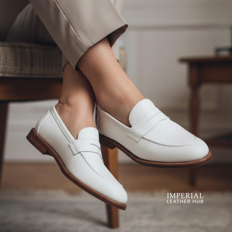 K&ouml;nnte beinhalten: Wei&szlig;e Penny-Loafer aus Leder mit braunen Sohlen. Die Schuhe haben ein klassisches Design mit einem Riemen &uuml;ber dem Spann. Das Bild zeigt die Schuhe an den F&uuml;&szlig;en einer Person, mit einer neutralfarbenen Hose.