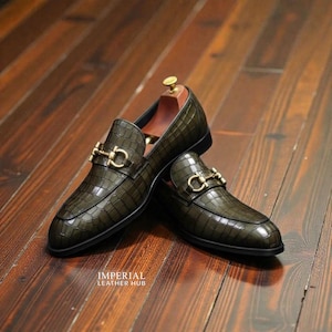 Könnte beinhalten: Ein Paar olivgrüne Leder-Loafer mit Krokodiltextur und goldfarbenen Metallakzenten. Die Schuhe haben ein klassisches Design. Sie werden auf einer Holzoberfläche präsentiert. Die Schuhe sind ca. 30 cm lang.