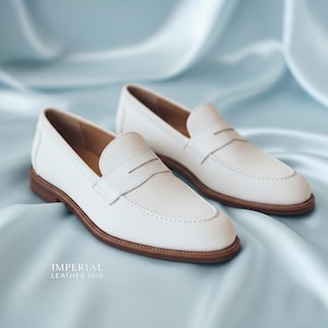 K&ouml;nnte beinhalten: Ein Paar wei&szlig;e Leder-Loafer mit Penny-Riemen-Design. Die Schuhe haben braune Sohlen und sind auf einem hellblauen Satinhintergrund abgebildet. Der Text "IMPERIAL LEATHER HUB" ist sichtbar.