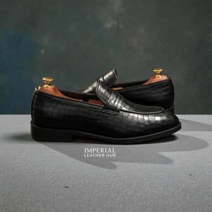 Pu&ograve; includere: Un paio di mocassini in pelle nera con una texture a pelle di coccodrillo. Le scarpe hanno un tacco basso e una punta arrotondata. Il testo "IMPERIAL LEATHER HUB" &egrave; stampato.