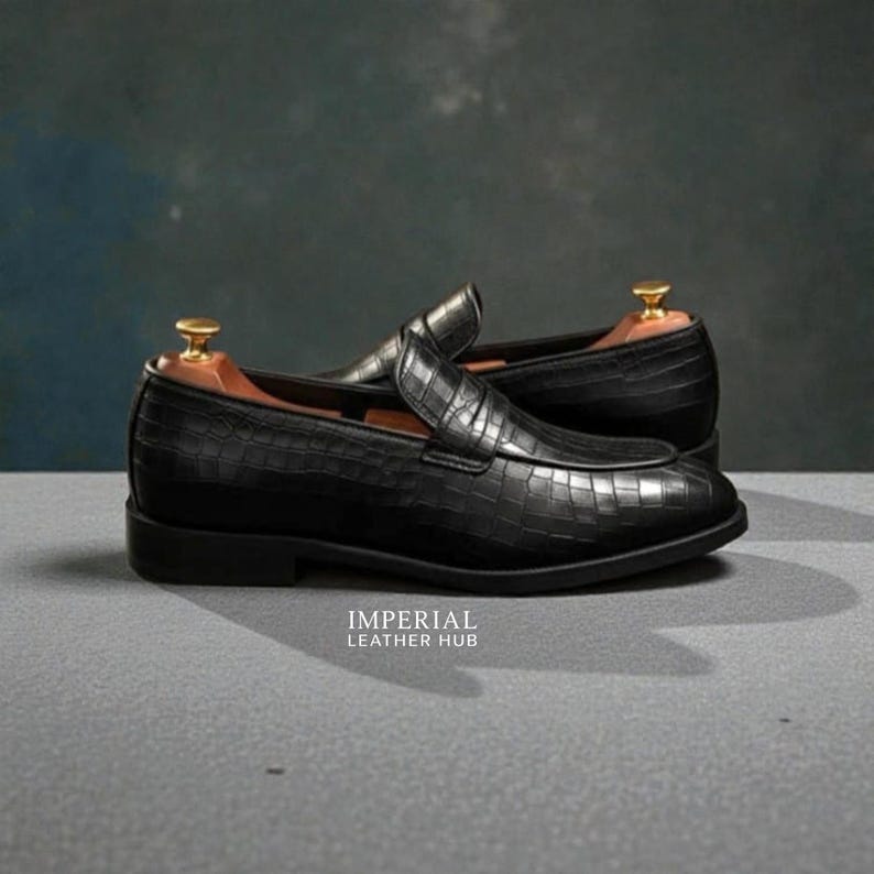 Pu&ograve; includere: Un paio di mocassini in pelle nera con una texture a pelle di coccodrillo. Le scarpe hanno un tacco basso e una punta arrotondata. Il testo "IMPERIAL LEATHER HUB" &egrave; stampato.
