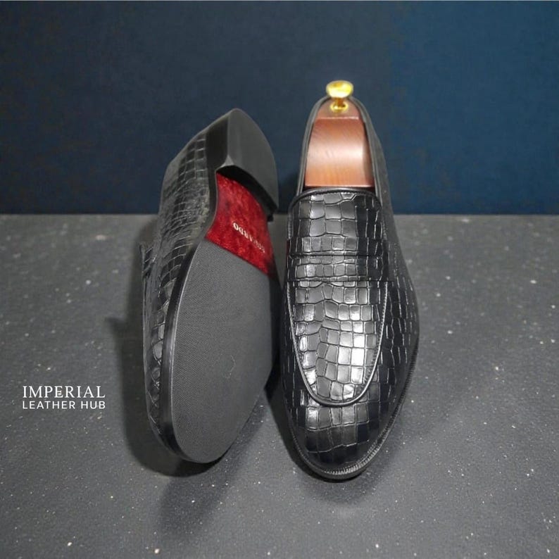 Pu&ograve; includere: Mocassini in pelle nera con texture a coccodrillo. Una scarpa &egrave; in posizione verticale, l'altra &egrave; capovolta, rivelando una suola rossa. Le scarpe sono esposte su una superficie scura.