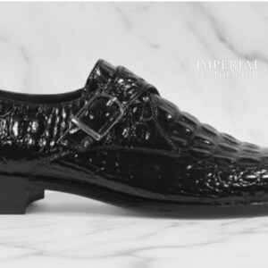 May include: A black leather shoe with a crocodile pattern. The shoe has a buckle closure and a low heel. The shoe is shiny and appears to be new. The shoe is a formal style.