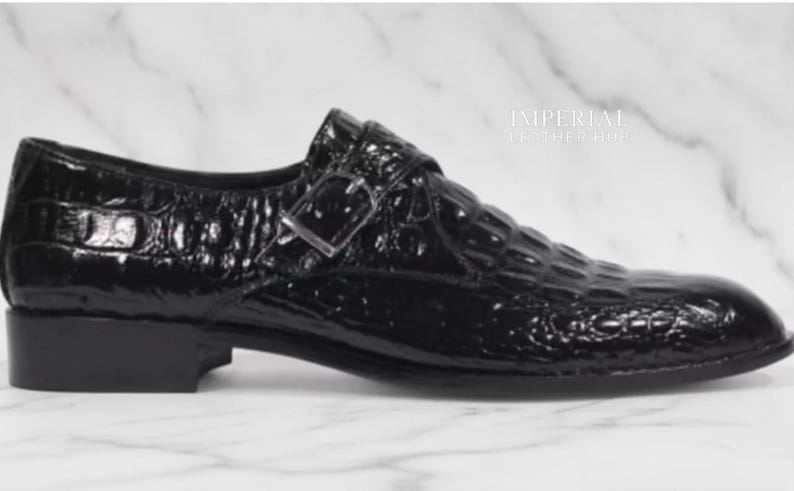 May include: A black leather shoe with a crocodile pattern. The shoe has a buckle closure and a low heel. The shoe is shiny and appears to be new. The shoe is a formal style.