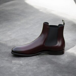 Pu&ograve; includere: Un singolo stivale Chelsea in pelle bordeaux lucida. Lo stivale presenta una punta arrotondata, pannelli laterali elastici e un tirante sul retro. La suola &egrave; nera e lo stivale &egrave; su uno sfondo grigio neutro. Il testo "IMPERIAL LEATHER HUB" &egrave; visibile.