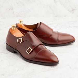 Peut inclure: Une paire de chaussures &agrave; boucle en cuir marron brillant, avec des boucles dor&eacute;es. Les chaussures sont pr&eacute;sent&eacute;es sur une surface en marbre, mettant en valeur leur design &eacute;l&eacute;gant et leur savoir-faire. Des embauchoirs en bois sont ins&eacute;r&eacute;s &agrave; l'int&eacute;rieur.