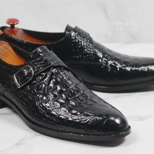 May include: A pair of black leather dress shoes with a crocodile skin texture. The shoes have a buckle closure and are displayed with wooden shoe trees. The shoes are on a white surface.